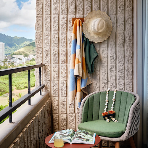 A cozy balcony features a green chair, side table with a drink and magazine, colorful towels, and a hat hanging on the wall, with a scenic view outside.