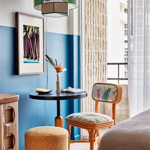 A cozy, modern room features a round table with chairs, vibrant decor, a hanging lamp, framed art, and a bed with beige bedding near an open window.