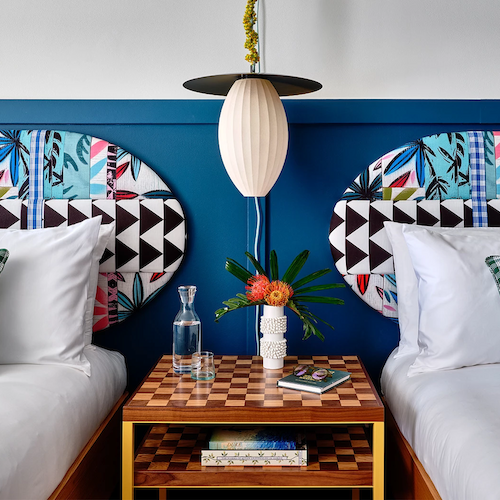 A well-decorated room with two beds featuring colorful headboards, checkered pillows, a shared checkered side table, a lamp, and decor items.