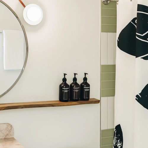 A bathroom with a round mirror, a towel, three bottles on a wooden shelf, and a shower curtain with a black and white abstract design.