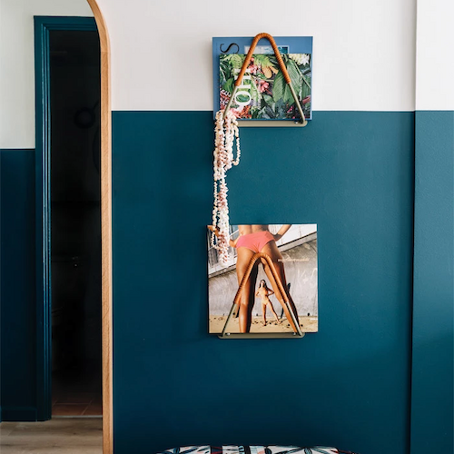 The image shows a room with a half teal, half white wall, decorated with two photos, a mirror, and a patterned cushioned seat beneath the decor.