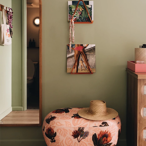 A room with a floral ottoman, a straw hat, a wall mirror, two hanging pictures, and a dresser with a box. The walls are painted light green.