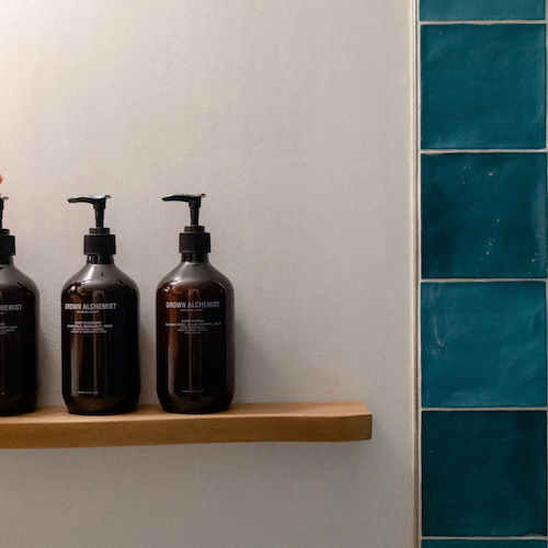 A hand reaches for one of three brown pump bottles on a wooden shelf against a white wall, next to a blue tiled wall.