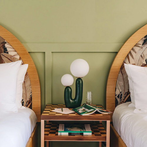 A bedroom with two beds featuring decorative headboards and green-checkered pillows, separated by a nightstand holding a cactus-shaped lamp and books.