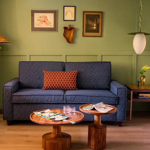 A cozy living space with a blue sofa, wooden tables, a bed nearby, and wall art. A lamp is in the corner. Magazines are on the tables.