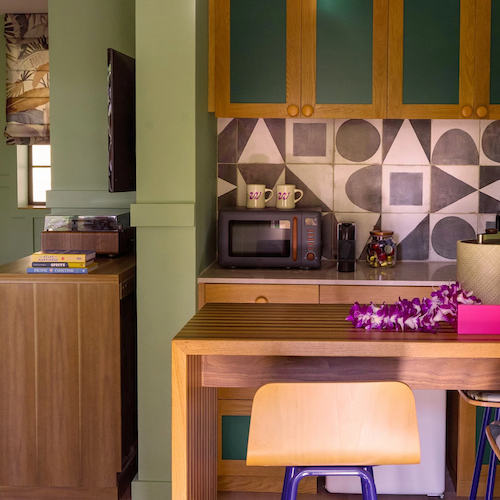 A cozy kitchenette with a microwave, cabinets, and geometric tiled backsplash. A small bar area and a living space featuring a bed and a wooden console.