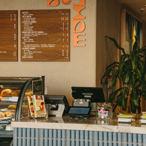 This image shows a café counter with a cash register, a plant, a menu on the wall, and a glass display case with food items.