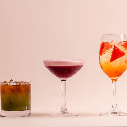 Three drinks are displayed: a green cocktail in a lowball glass, a purple cocktail in a coupe glass, and a citrus cocktail in a wine glass.