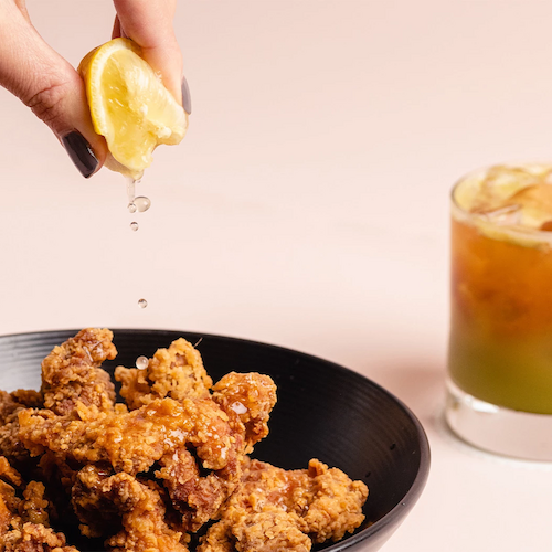 A hand squeezes a lemon over a bowl of fried chicken pieces, with a glass of a mixed drink in the background ending the sentence.