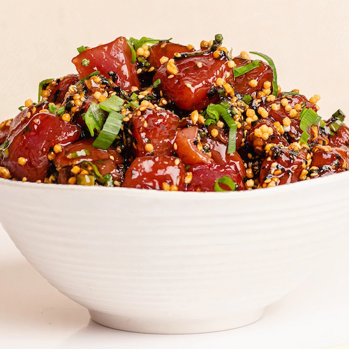 A white bowl filled with a colorful poke dish, featuring diced raw fish, sesame seeds, green onions, and other fresh ingredients.