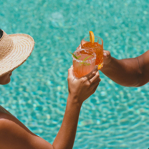 Two people toasting with colorful drinks by a pool, one wearing a wide-brimmed hat and the other shirtless.