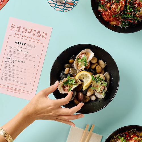 A person reaches for a bowl of oysters with lemon slices and garnishes; nearby are sushi bowls, a menu with 