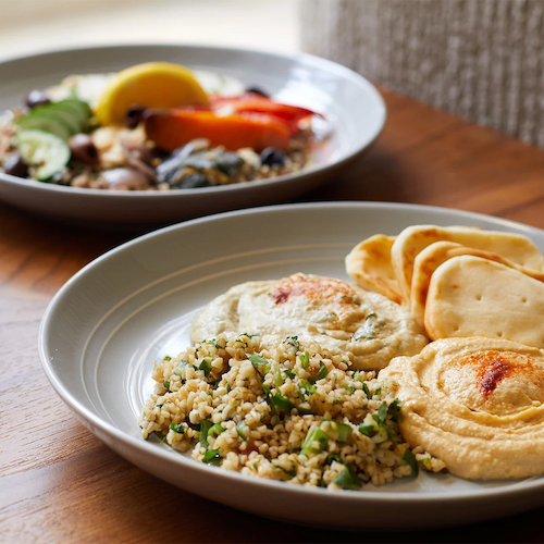 The image shows two plates of food. The closer plate contains hummus, pita bread, and tabbouleh salad. The second plate has roasted vegetables.