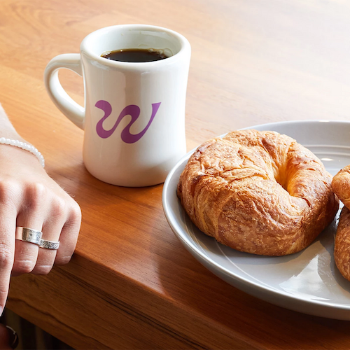 The image shows a hand wearing rings and a bracelet, a white coffee mug with a purple 