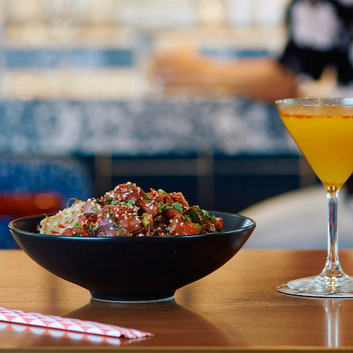 A bowl of food, likely a salad or poke, is next to a yellow cocktail in a martini glass, with a red-striped straw on the table.