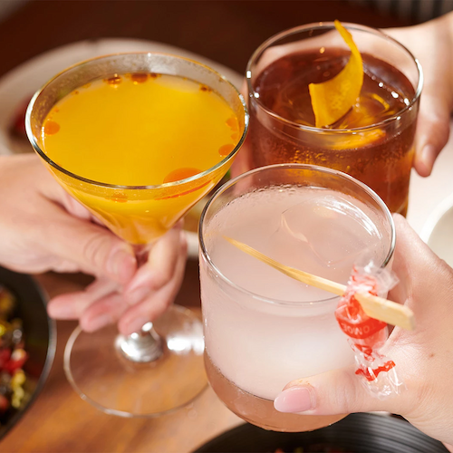 Three people clinking their glasses of cocktails together, with food visible on the table.