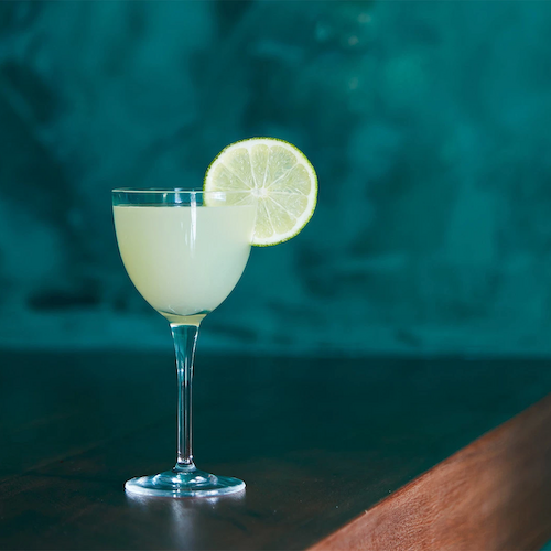 A cocktail glass with a green drink and a lime slice garnish on a wooden table against a green background, in a somewhat dimly lit environment.