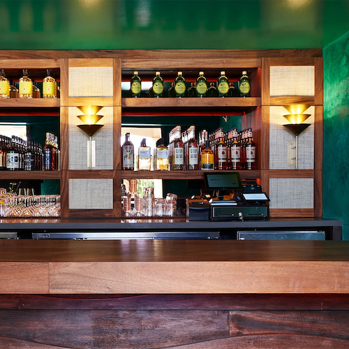 A bar area featuring wooden shelves stocked with various bottles of liquor, a cash register, and warm lighting fixtures.