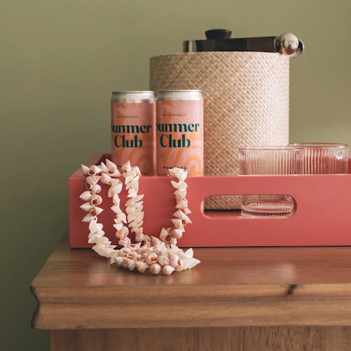 A wooden surface holds a pink tray with two cans, a woven ice bucket, drinkware, and seashell necklaces, set against a green wall.