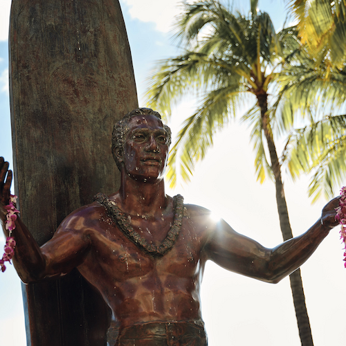 A statue of a shirtless figure holding leis, with palm trees and a surfboard in the background, bathed in sunlight.