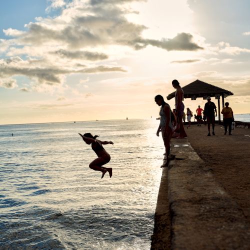 People are on a pier, with one person mid-jump into the ocean against a sunset sky. Others stand nearby, silhouetted by the sunlight.