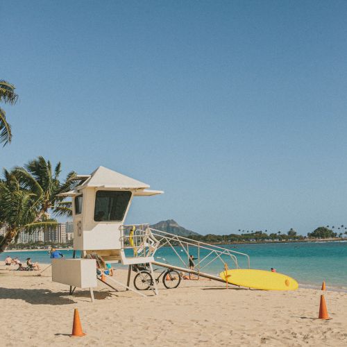 A sandy beach with a lifeguard tower, palm trees, and a yellow kayak by the turquoise water under a clear blue sky.