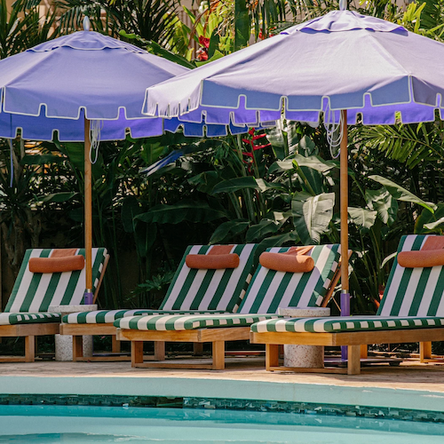 The image shows a pool area with striped lounge chairs and purple umbrellas, surrounded by lush greenery and tropical plants.