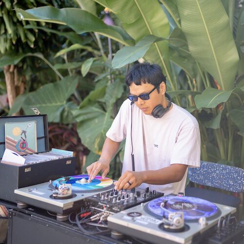 A person wearing sunglasses and headphones is DJing on a turntable setup outdoors, surrounded by large green plants.