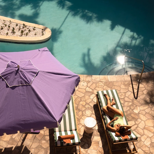 A person is lounging on a striped chair under a purple umbrella beside a pool, with another empty chair and a small table nearby.