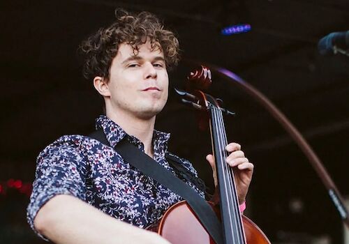 A person with curly hair plays a cello on stage, wearing a patterned shirt and a strap over their shoulder, with stage lights in the background.