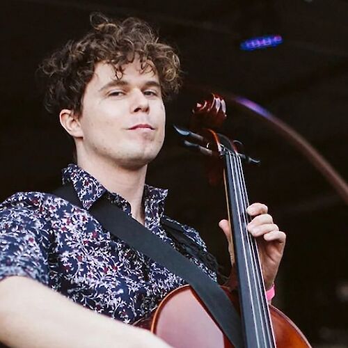 A person with curly hair plays a cello on stage, wearing a patterned shirt and a strap over their shoulder, with stage lights in the background.