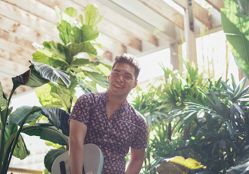 A man is standing and smiling in front of lush green plants and trees, holding a guitar. The setting appears bright and natural, with sunlight streaming in.