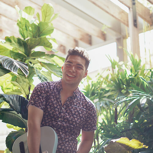 A man is standing and smiling in front of lush green plants and trees, holding a guitar. The setting appears bright and natural, with sunlight streaming in.