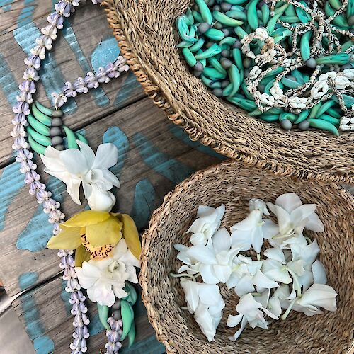 The image shows two wicker baskets with turquoise and white beads, and a floral necklace made of white, yellow, and green flowers and beads.