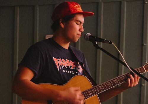 A person wearing a red cap and dark shirt is playing an acoustic guitar and singing into a microphone. The background appears indoors with dim lighting.