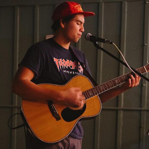 A person wearing a red cap and dark shirt is playing an acoustic guitar and singing into a microphone. The background appears indoors with dim lighting.