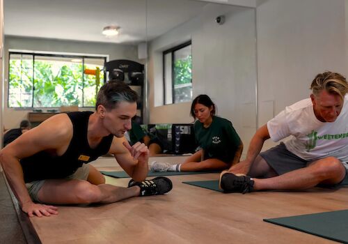 People are engaging in a stretching exercise in a well-lit room with large windows and exercise equipment visible in the background.