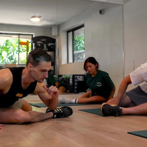 People are engaging in a stretching exercise in a well-lit room with large windows and exercise equipment visible in the background.