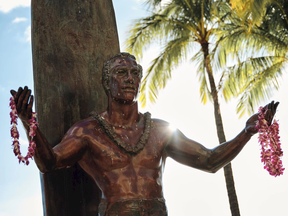 The image features a bronze statue of a surfer holding leis, with palm trees and a surfboard in the background, captured in bright sunlight.