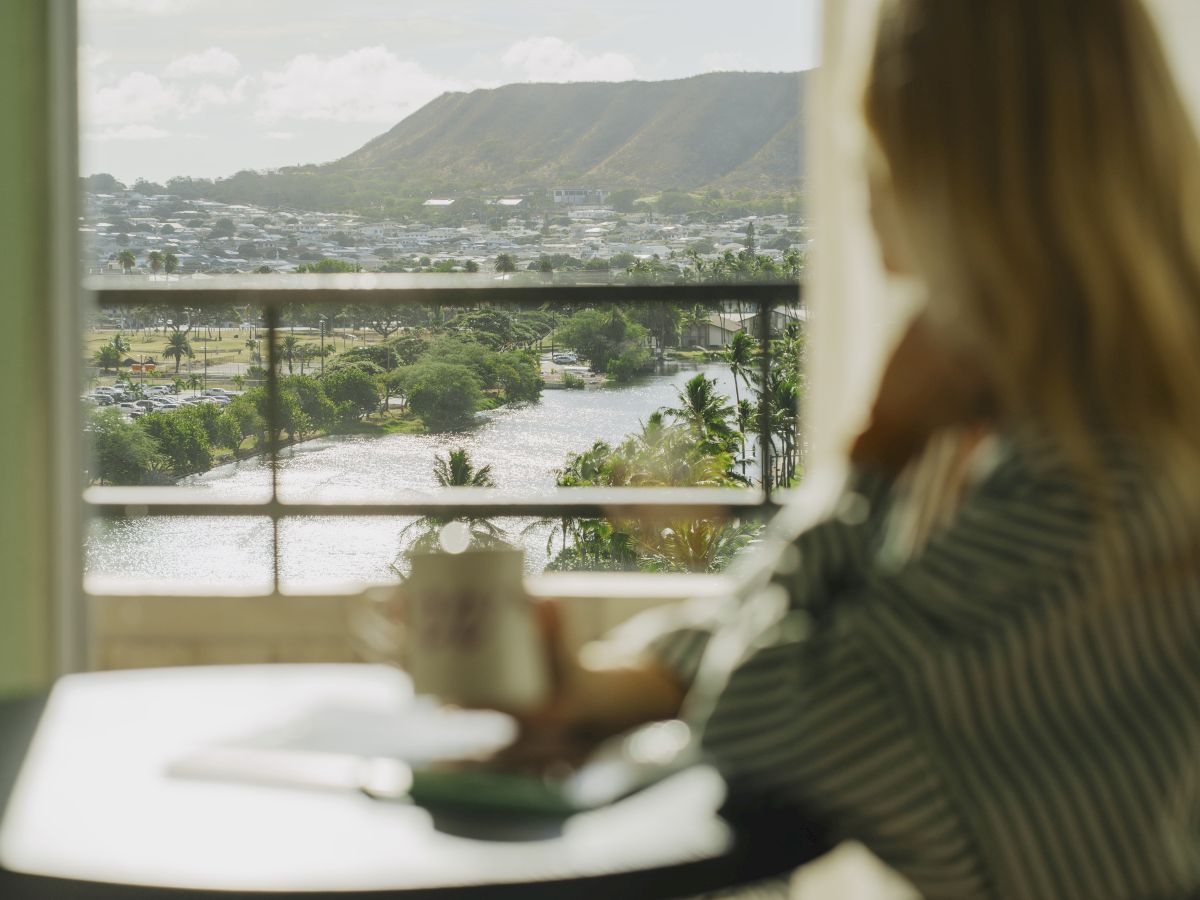 Une personne est assise &agrave; une table &agrave; l'int&eacute;rieur, regardant par la fen&ecirc;tre une rivi&egrave;re et des collines au loin, floues au premier plan.