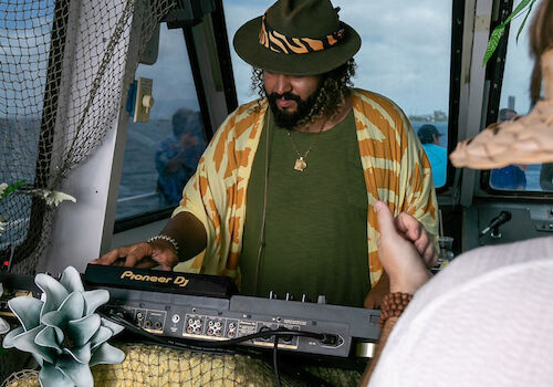 A person wearing a hat and patterned jacket is playing a keyboard or mixer in an outdoor setting, possibly on a boat, surrounded by decorations.