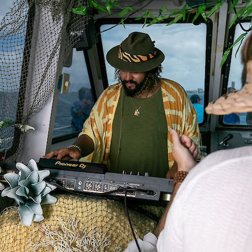 A person wearing a hat and patterned jacket is playing a keyboard or mixer in an outdoor setting, possibly on a boat, surrounded by decorations.