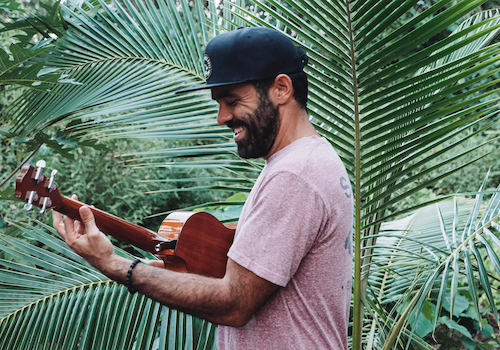 A man wearing a black cap, smiling, and playing a guitar stands among lush green palm leaves.