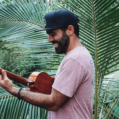 A man wearing a black cap, smiling, and playing a guitar stands among lush green palm leaves.