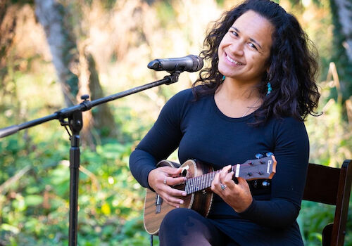 A woman sits on a chair outdoors, playing a ukulele and smiling, with a microphone in front of her, surrounded by greenery.