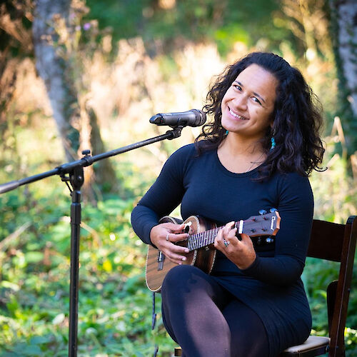 A woman sits on a chair outdoors, playing a ukulele and smiling, with a microphone in front of her, surrounded by greenery.