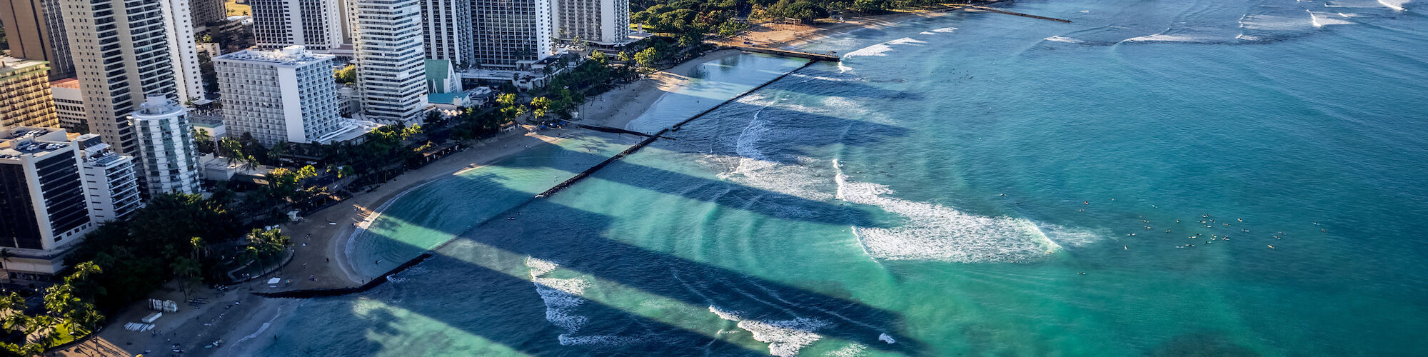 A coastal cityscape with high-rise buildings, turquoise waters, surfers, and a mountain in the background under a clear sky, peaceful and inviting.