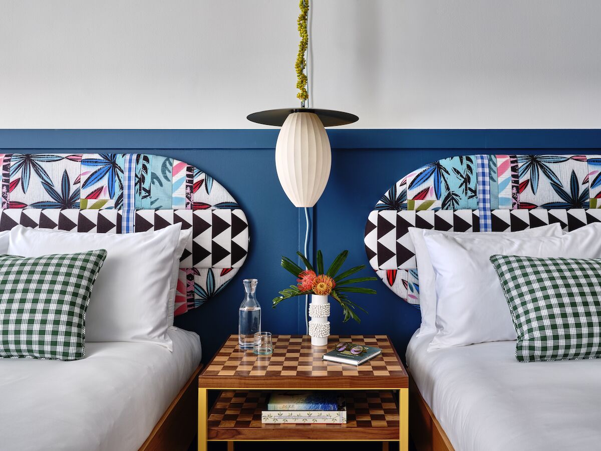 The image shows two beds with colorful headboards, a patterned lamp, a checkered pillow, and a small table with items on it between the beds.