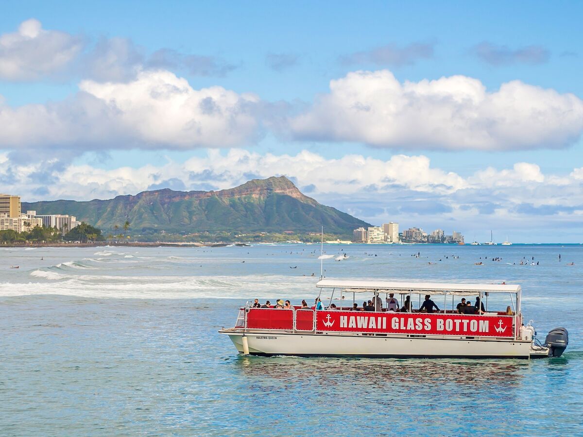 A glass-bottom boat on the ocean with a mountainous backdrop, likely near a coast, under a partly cloudy sky.