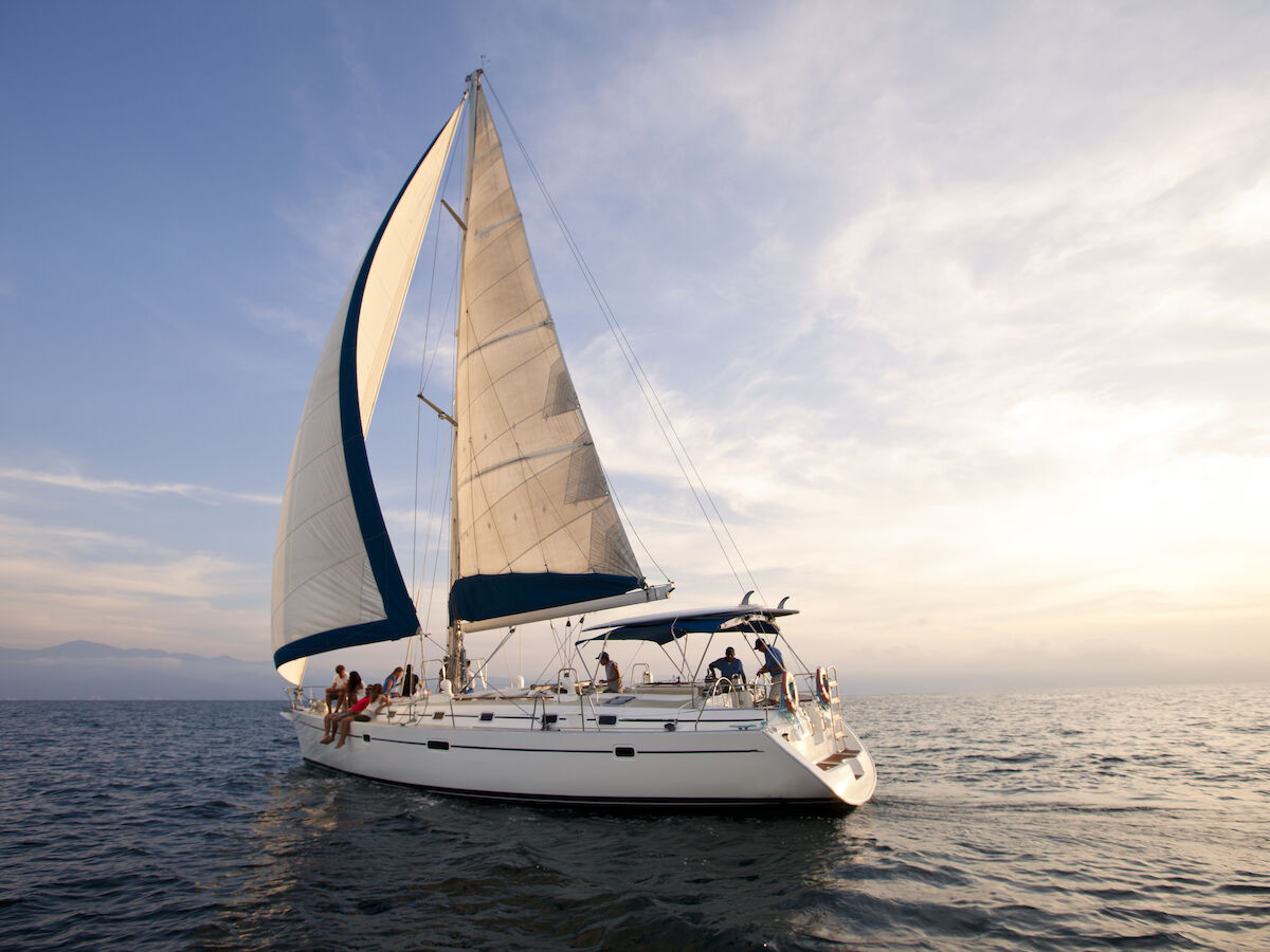 A white sailboat with its sails up on a calm body of water, with people onboard and a picturesque sky in the background.